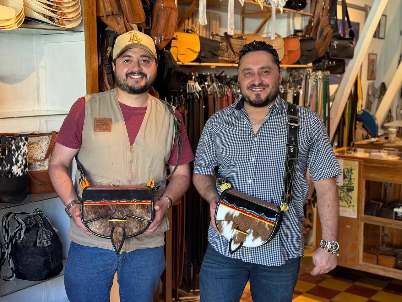 Dos hombres sonrientes, uno con gorra y chaleco y otro con camisa a cuadros, sostienen carrieles de cuero dentro de un taller con correas, bolsos y sombreros colgados.