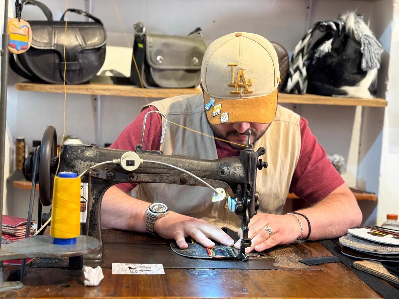 Un hombre con gorra cose una pieza de cuero en una máquina de coser antigua, con un carrete de hilo amarillo y bolsos de cuero en los estantes de fondo.