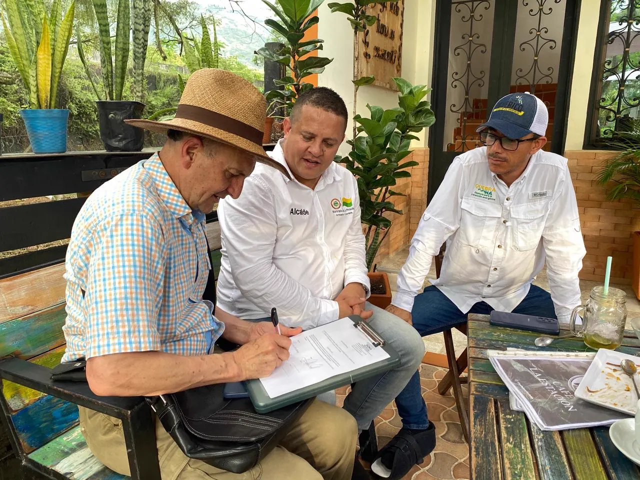 Tres hombres sentados alrededor de una mesa; el de sombrero firma un documento sobre una tabla con clip mientras los otros dos lo observan, rodeados de plantas.