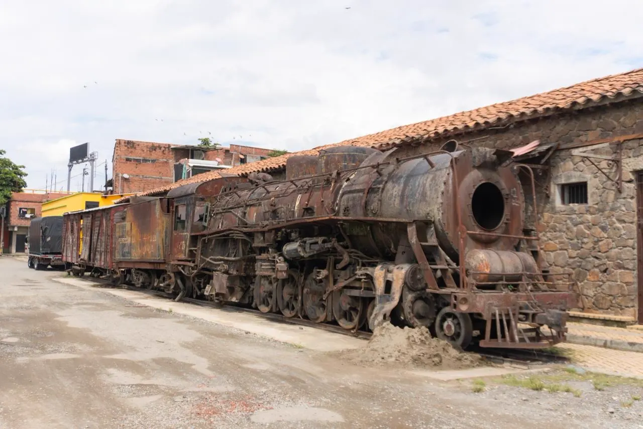 Una locomotora a vapor oxidada con vagones de carga, detenida junto a la fachada de piedra y techo de tejas de la antigua estación.