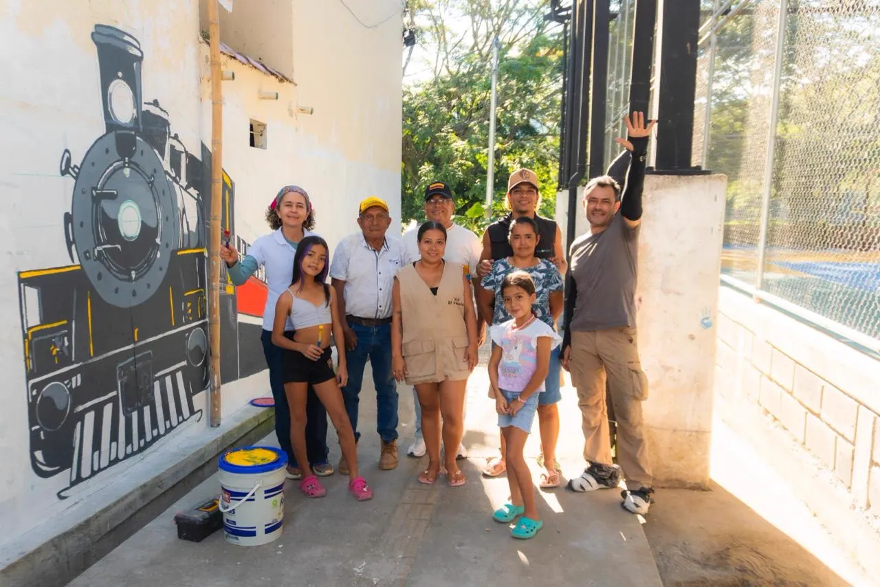 Un grupo de adultos y niñas posa en un pasillo junto a un muro con una locomotora pintada en blanco y negro; en el piso hay baldes de pintura.