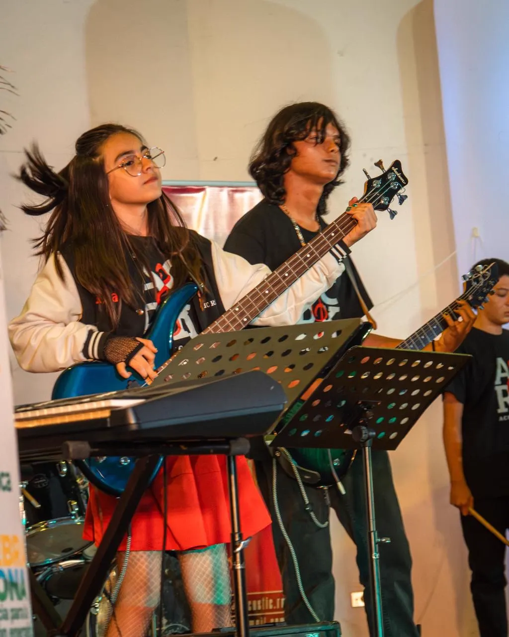 Una joven con gafas toca un bajo eléctrico azul junto a un joven de cabello largo con guitarra, frente a atriles de partituras.