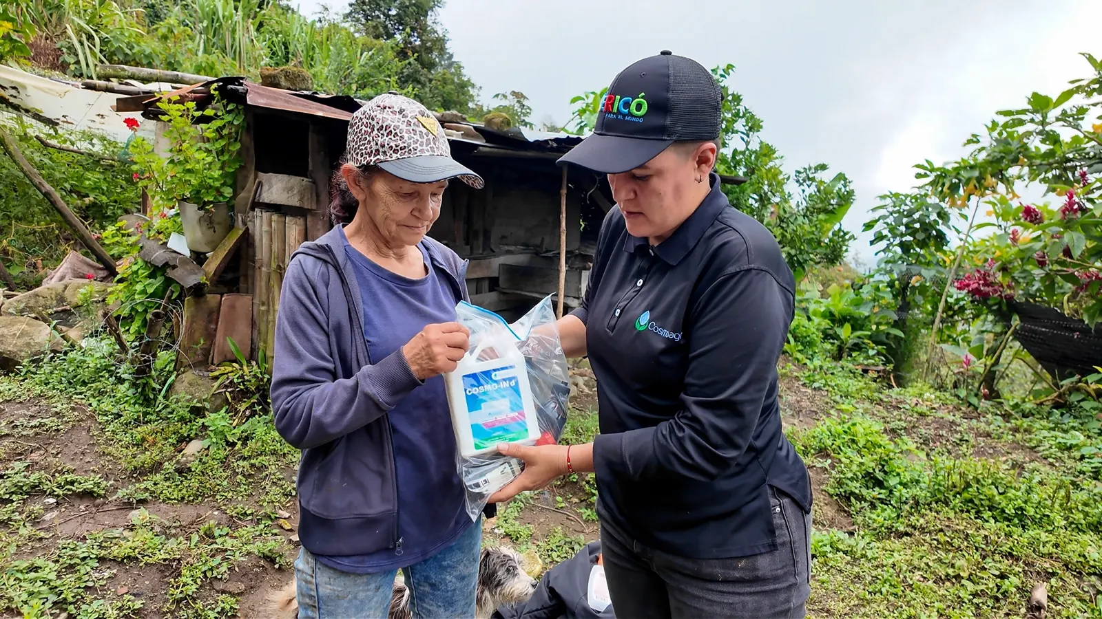 Una mujer mayor con gorra estampada recibe una bolsa con un envase de insumos de manos de una mujer con gorra y camisa negras, frente a una casa rural rodeada de vegetación.