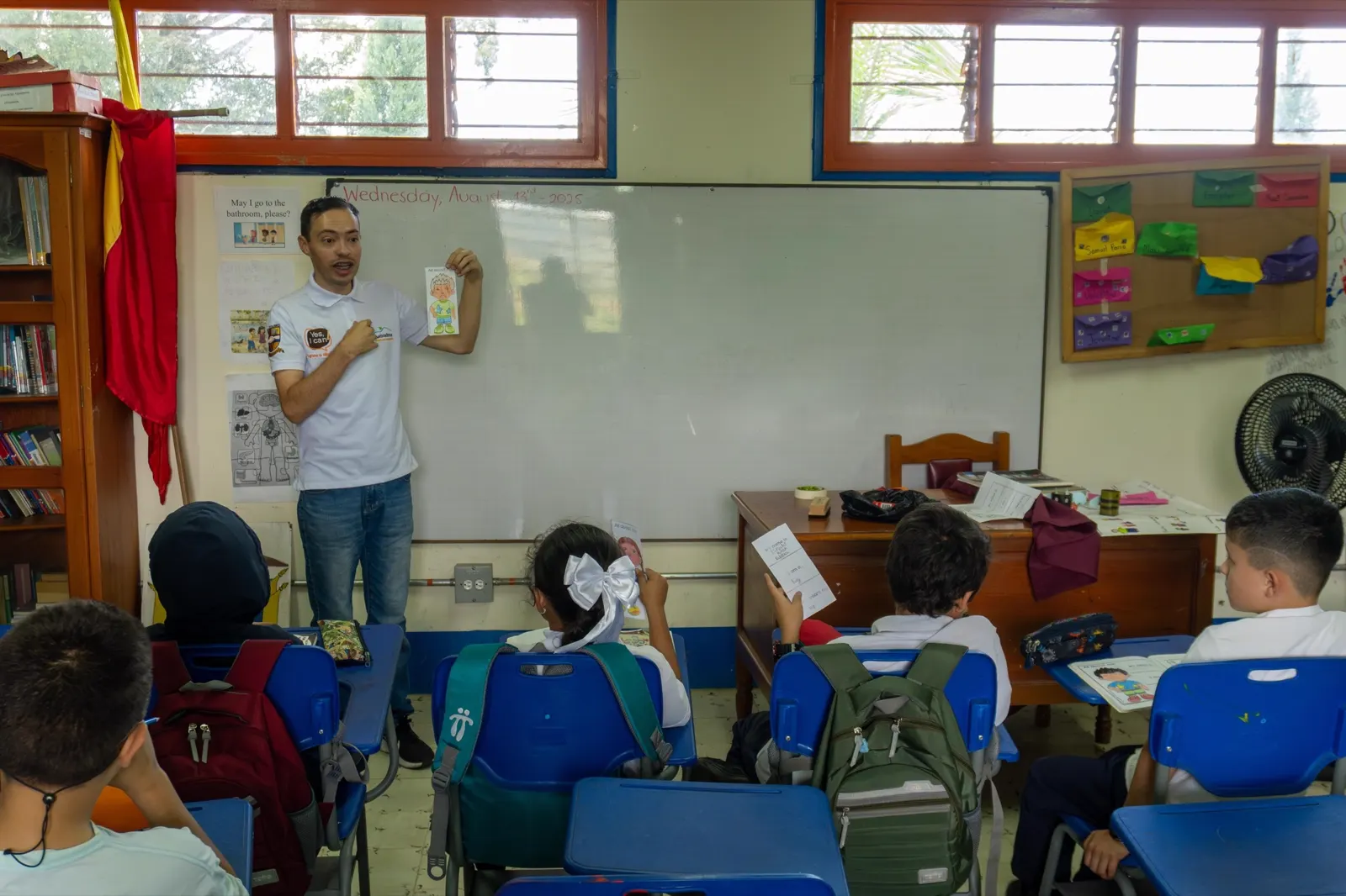 Un profesor con camiseta blanca de pie frente al tablero de un salón de clases, hablando a un grupo de niños sentados en pupitres azules.