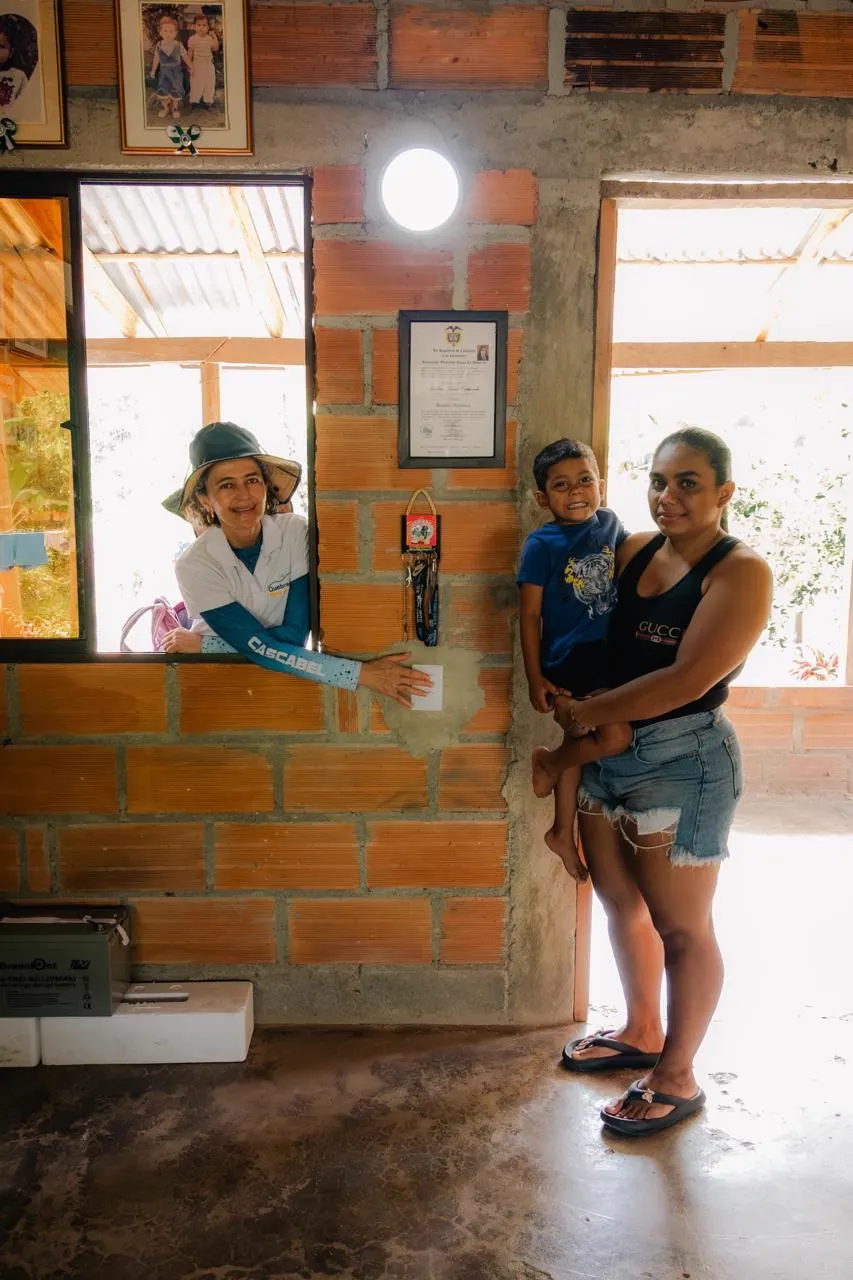 Una mujer con sombrero y camiseta de Quebradona se asoma por la ventana de una casa de ladrillo y señala un interruptor en la pared; a su lado, una mujer carga a un niño pequeño. En el techo se ve un bombillo encendido.