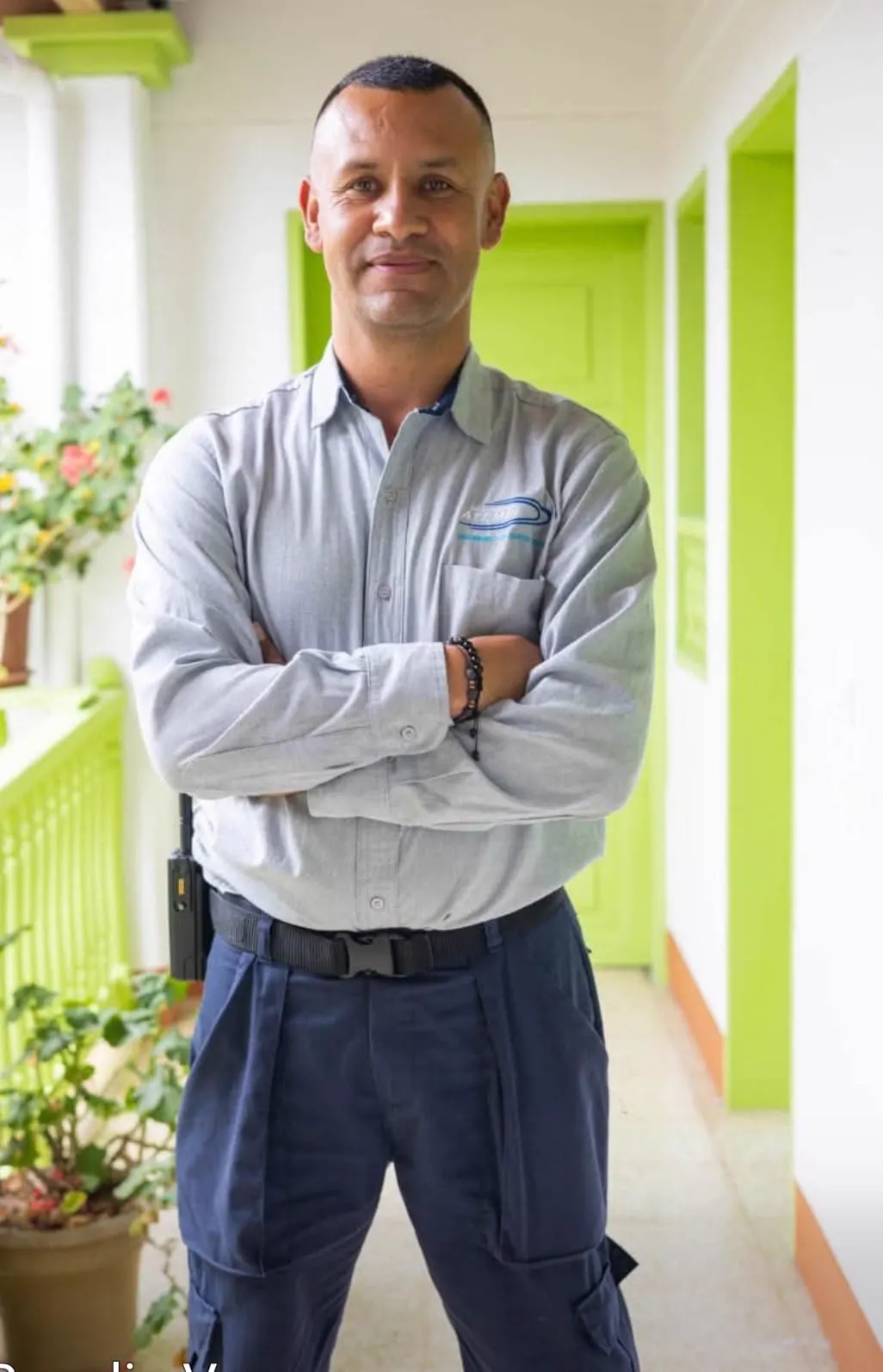 Retrato de un hombre con camisa gris de trabajo y los brazos cruzados, de pie en un corredor de paredes blancas y puertas verdes.