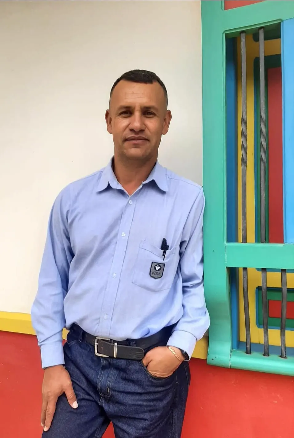 Un hombre con camisa azul clara y pantalón oscuro posa recostado junto a una ventana de madera pintada de colores.