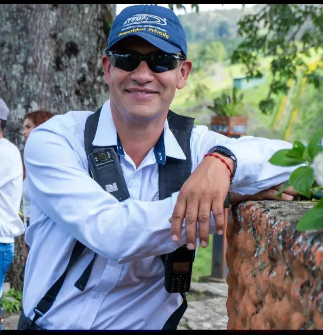 Un hombre sonriente con gorra azul, gafas oscuras y camisa clara con arnés, apoyado en un muro al aire libre.