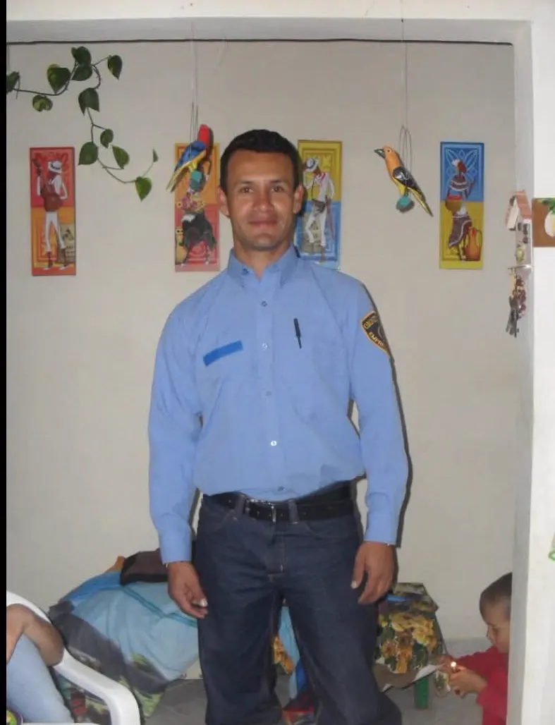 Un hombre con uniforme oscuro con franjas reflectivas y radio, sentado en un muro de ladrillo junto a la vegetación.