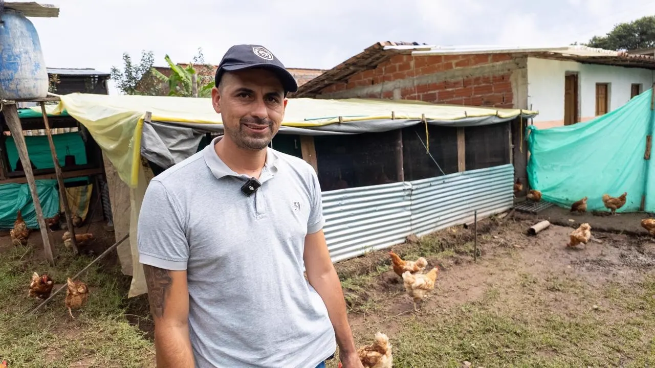 Un productor con gorra y camiseta gris posa frente a un corral de gallinas cubierto con lonas y láminas de zinc, junto a una casa de ladrillo.