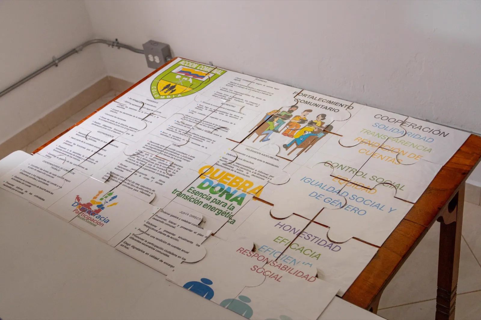 Un rompecabezas gigante armado sobre una mesa, con el logo de Quebradona y palabras como fortalecimiento comunitario, control social, igualdad social y de género, honestidad, eficacia y responsabilidad social.