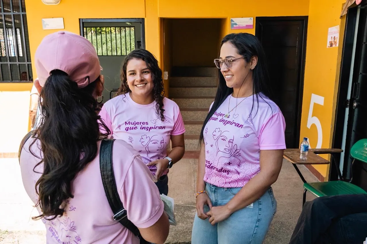 Dos mujeres sonríen con camisetas rosadas estampadas con el lema «Mujeres que inspiran», frente a un edificio de paredes amarillas; de espaldas, otra mujer con gorra rosada conversa con ellas.