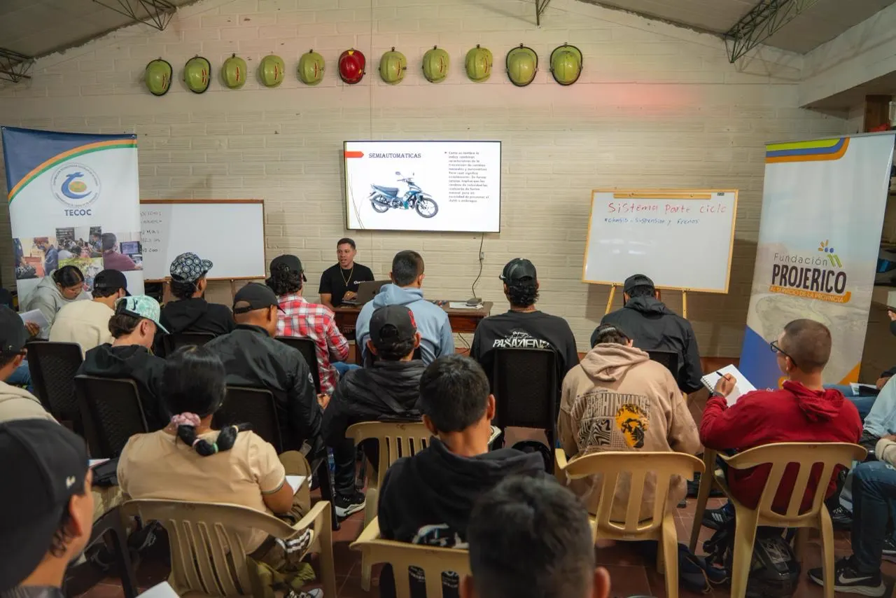 Participantes sentados en sillas plásticas, vistos de espaldas, atienden a un instructor que proyecta en pantalla la imagen de una motocicleta, en un salón con pendones de Tecoc y Fundación Projericó.