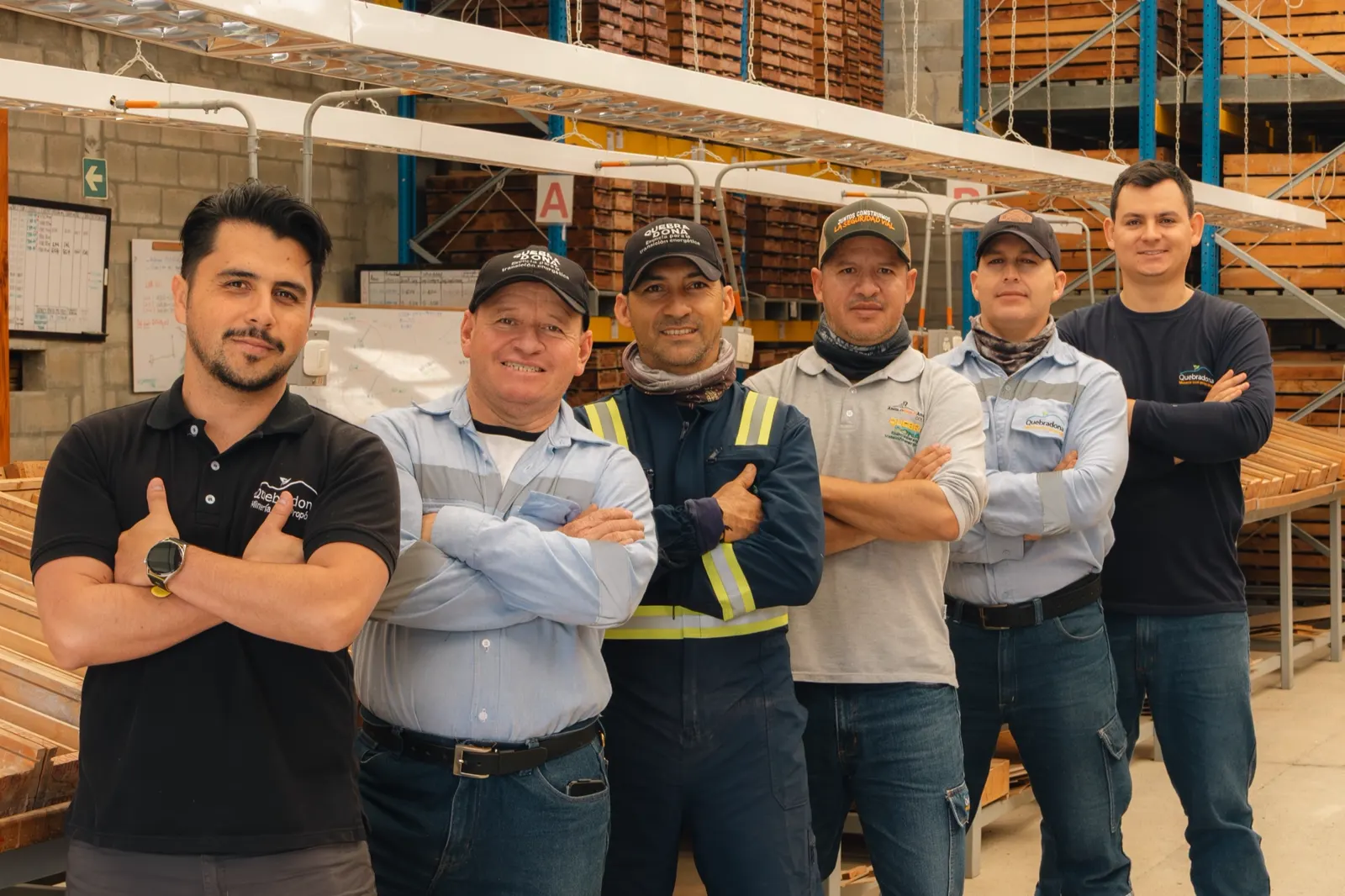 Seis hombres con los brazos cruzados posan sonrientes en una bodega con estanterías de cajas de muestras; varios llevan gorra y camisa de trabajo, uno con chaleco reflectivo.