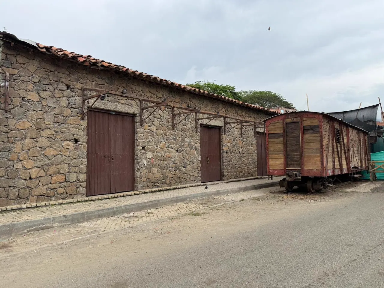 Fachada de piedra de la antigua estación del ferrocarril con puertas de madera y, a la derecha, un vagón de tren de madera y estructura roja junto a un cerramiento de tela verde.