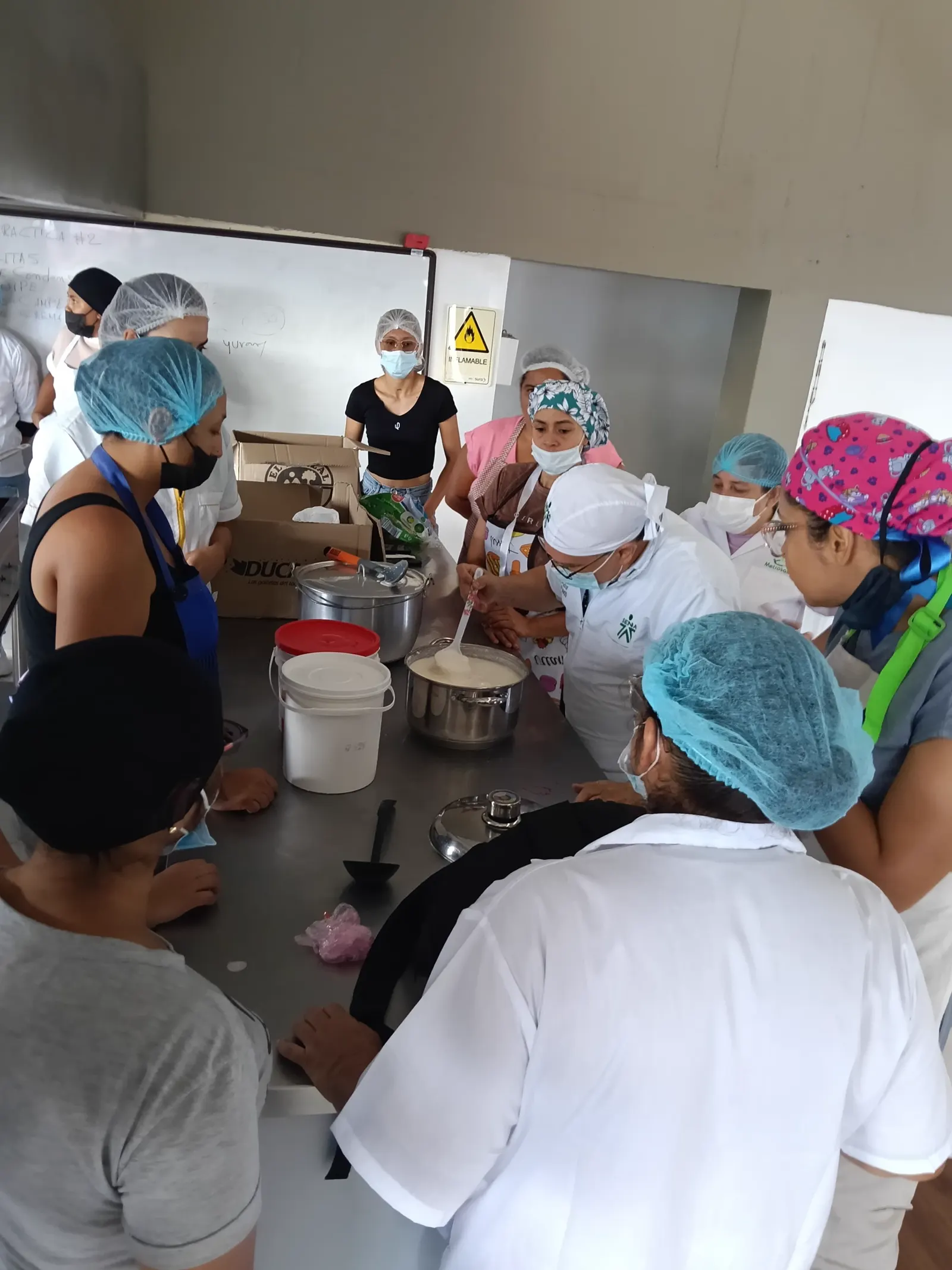 Participantes con gorros y tapabocas alrededor de una mesa de acero con ollas y baldes, mientras una mujer revuelve una preparación láctea.