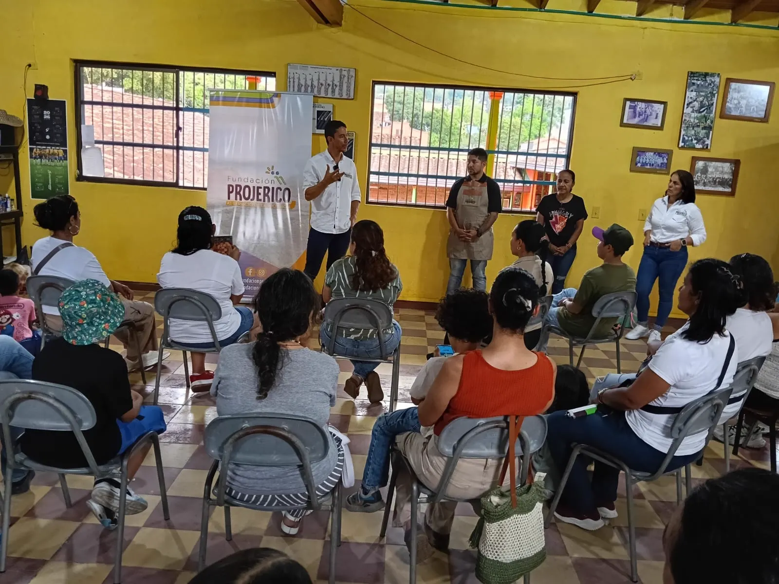 Personas sentadas en círculo escuchan a un hombre de camisa blanca que habla junto a un pendón de la Fundación Projericó, en un salón de paredes amarillas.