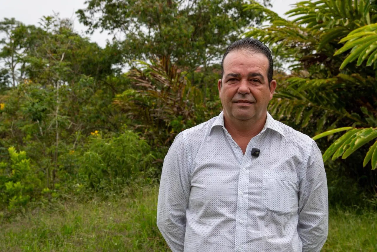 Retrato de Mario Sierra, hombre con camisa blanca de cuadros, de pie frente a árboles y vegetación.
