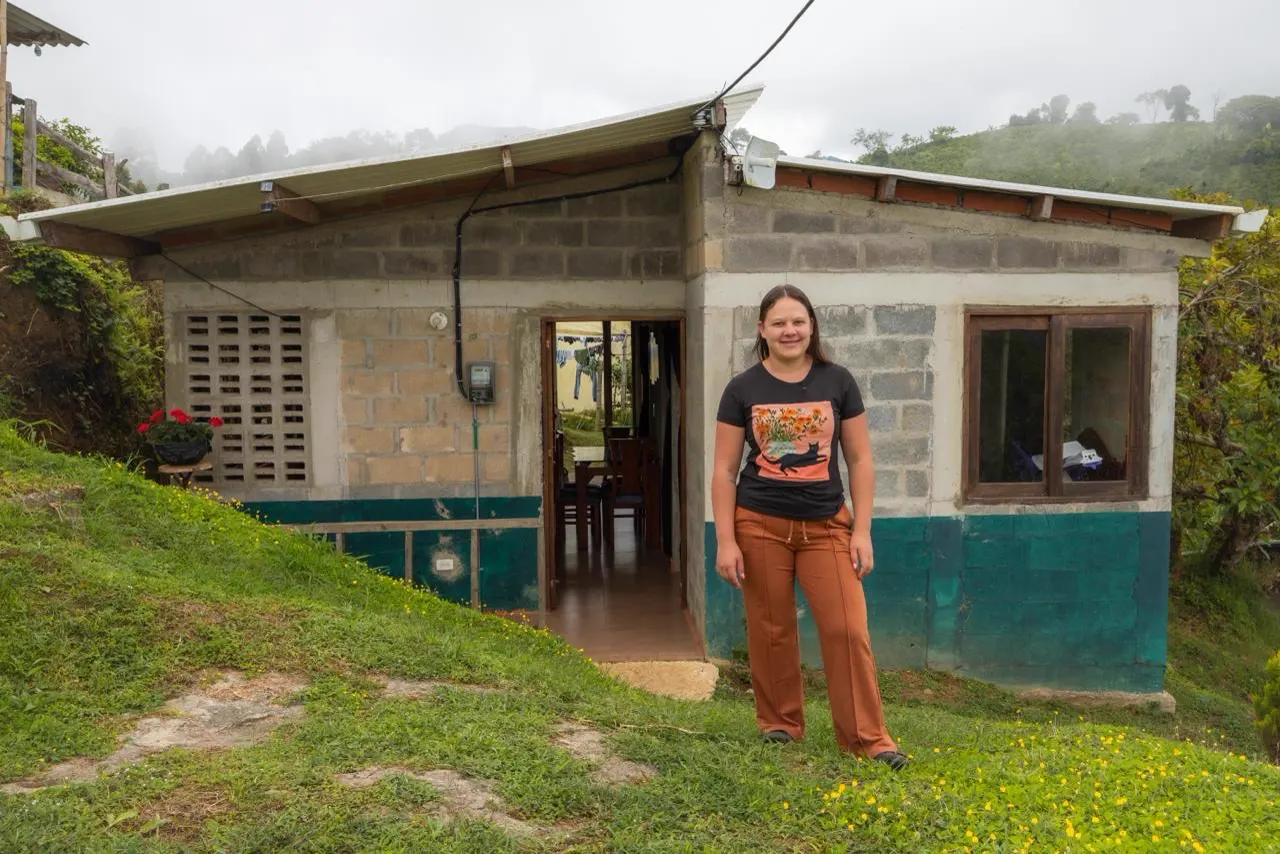 Una joven de pie sobre el pasto frente a la puerta de una casa rural de bloque con techo de lámina, en una ladera con neblina.