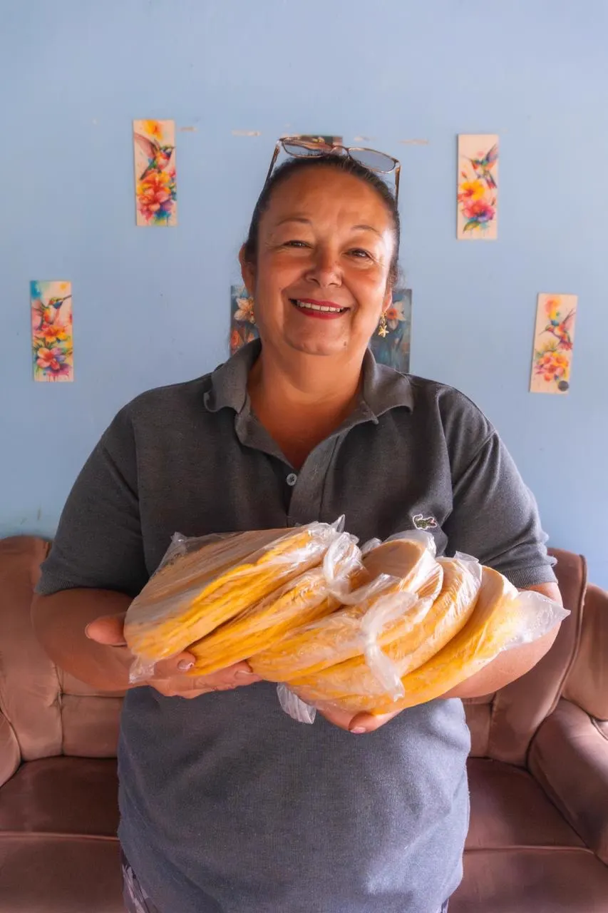 Dora Cano sonríe mientras sostiene varios paquetes de arepas envueltas en plástico, frente a una pared azul con cuadros de flores y colibríes.