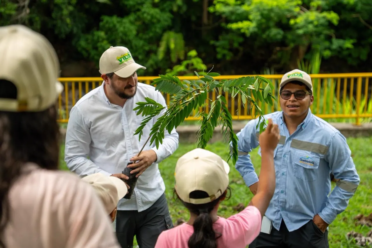 Dos hombres con gorras beige explican a un grupo de niños con gorras cómo sembrar una plántula de árbol que uno de ellos sostiene, en un prado junto a una baranda amarilla.