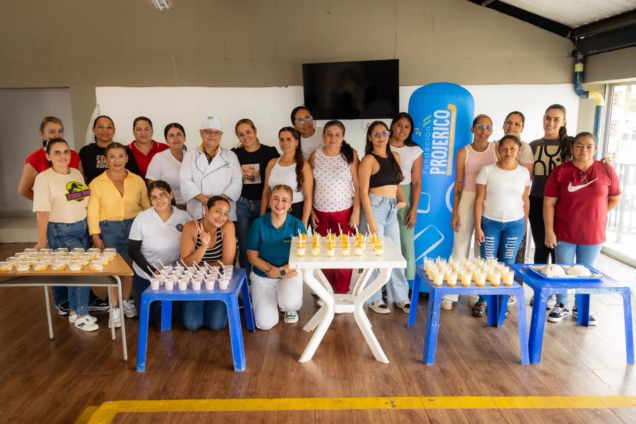 Grupo de unas veinte mujeres y un instructor de bata blanca posan en un salón junto a mesas con vasos de postres y un inflable azul de ProJericó.