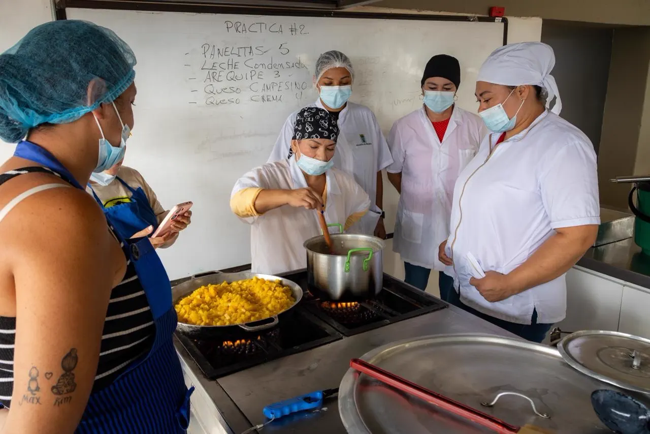 Mujeres con gorros, tapabocas y batas blancas cocinan alrededor de una estufa industrial con ollas, frente a un tablero con la lista de una práctica.