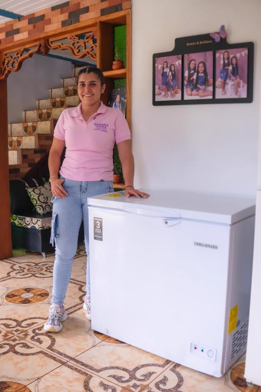 Sandra Cruz, de camiseta rosada, posa sonriente con la mano sobre un congelador horizontal blanco en la sala de una casa.