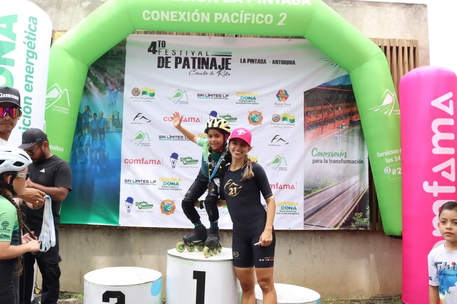 Una patinadora con gorra rosada y una niña con casco que saluda posan sobre el podio frente a un pendón con logos de patrocinadores, bajo un arco inflable verde.