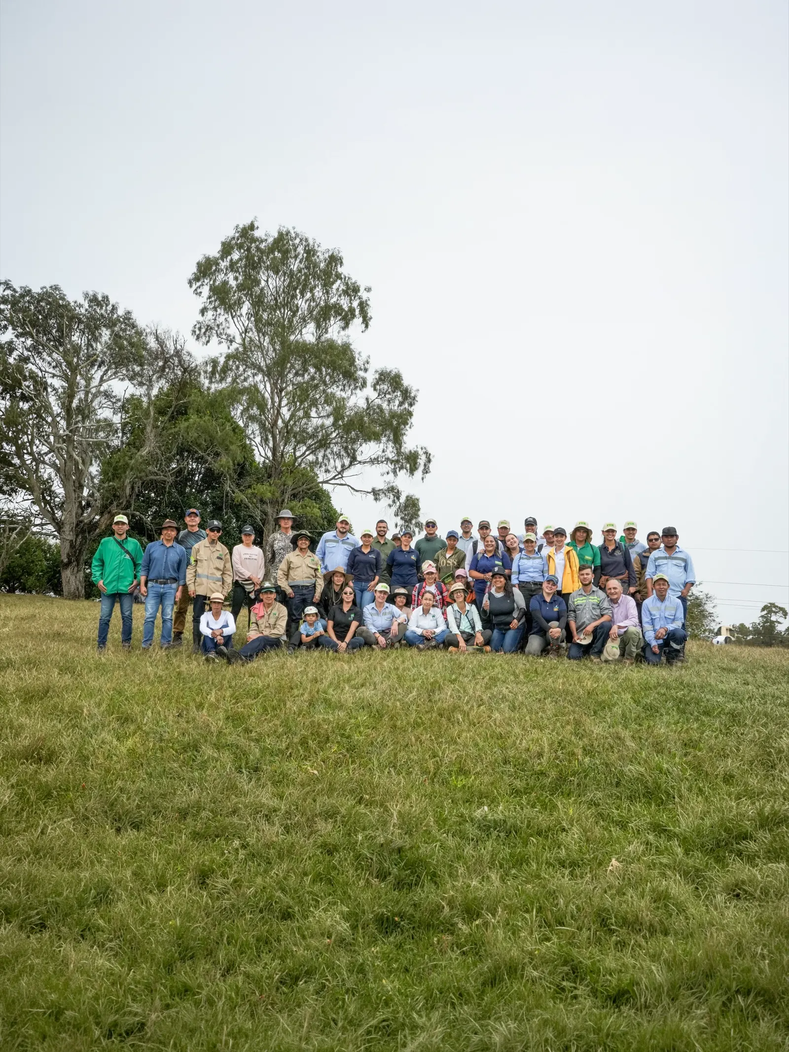Grupo numeroso de personas posa de pie y en cuclillas sobre un potrero verde, junto a árboles altos, bajo un cielo nublado.