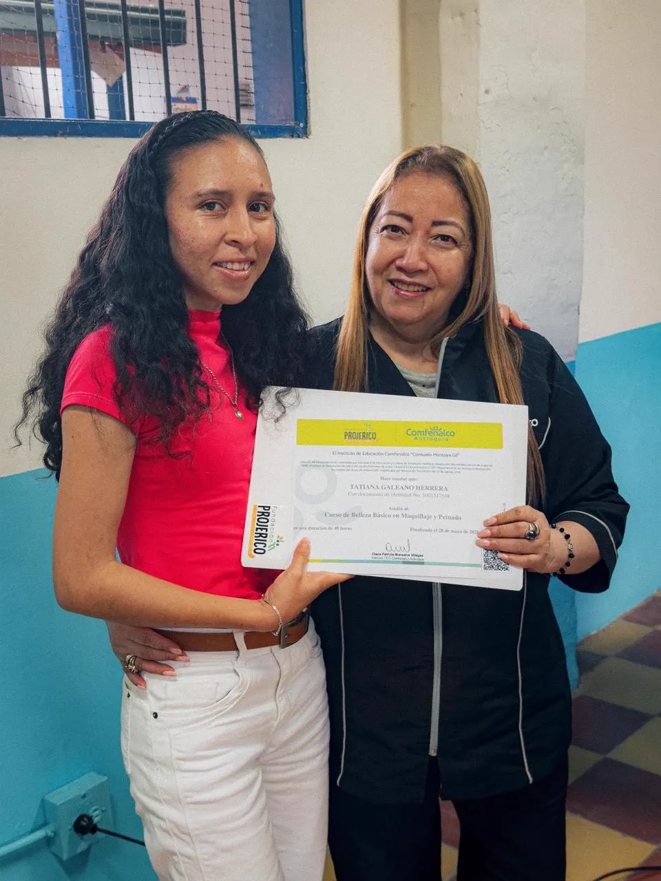 Una joven de blusa roja y una mujer de chaqueta oscura sostienen un certificado junto a una pared azul y blanca.