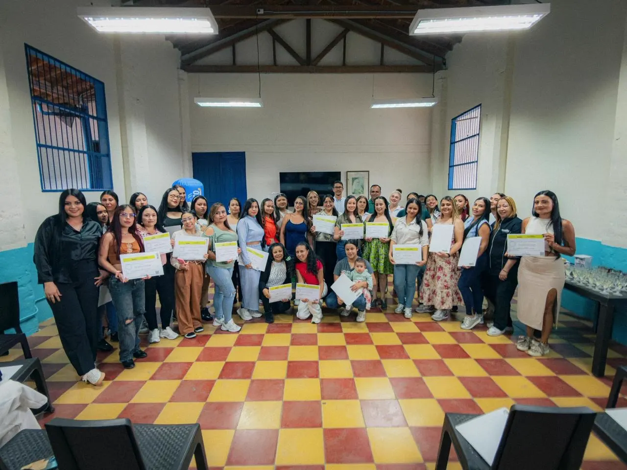 Decenas de mujeres y algunos hombres posan con sus certificados en un salón de piso ajedrezado amarillo y rojo, con paredes blancas y azules.