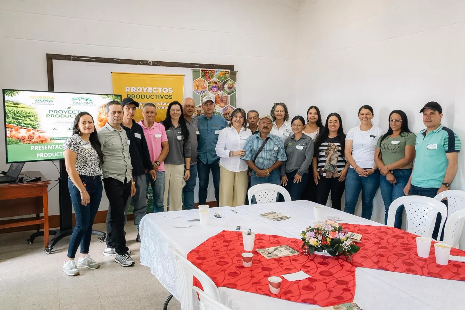 Un grupo de hombres y mujeres posa de pie en un salón, junto a una mesa con mantel rojo y flores, frente a una pantalla y un cartel de Proyectos Productivos.