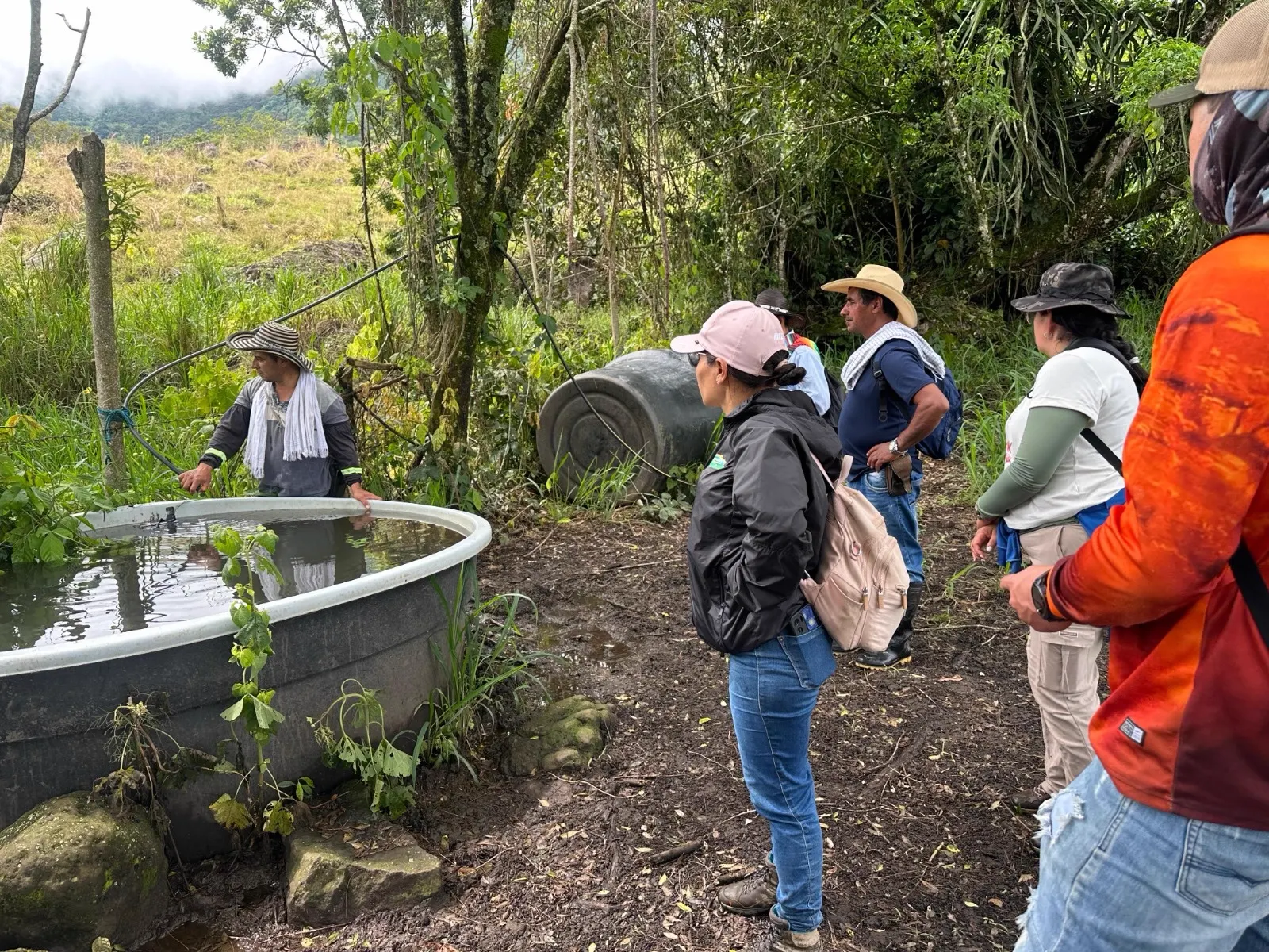 Varias personas con sombreros y gorras de pie alrededor de un tanque circular de agua en un terreno con árboles.