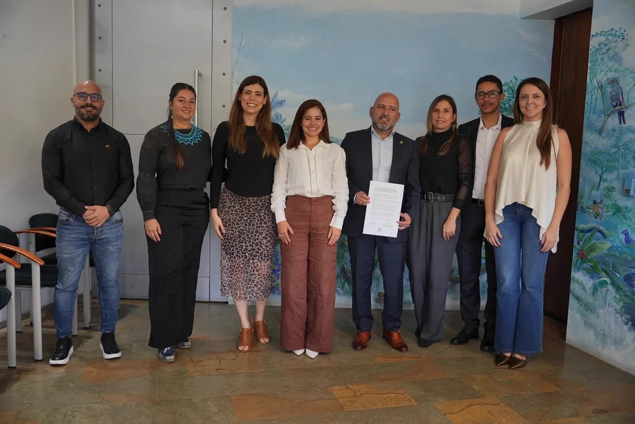 Grupo de nueve personas posa de pie en una sala con un mural de vegetación; un hombre en el centro sostiene el documento firmado.