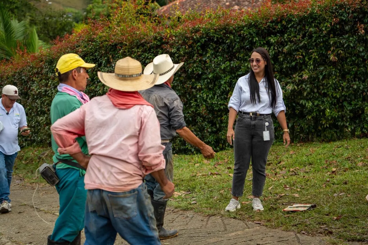 Varios campesinos con sombreros y gorras conversan en un camino veredal con una mujer de camisa clara y carné, frente a un seto verde.