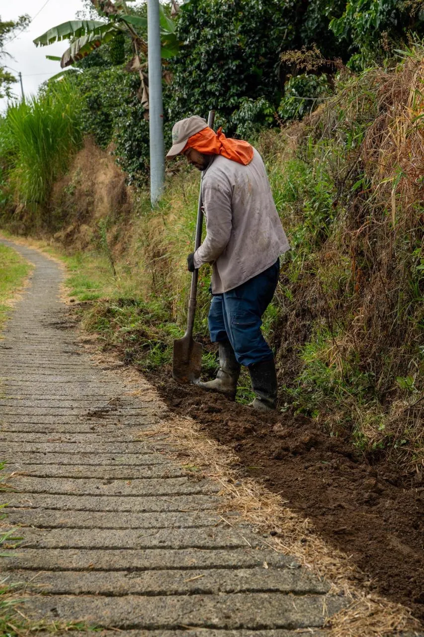 Un hombre con pañoleta naranja y gorra cava con una pala junto a un camino de concreto, al borde de un talud con vegetación.
