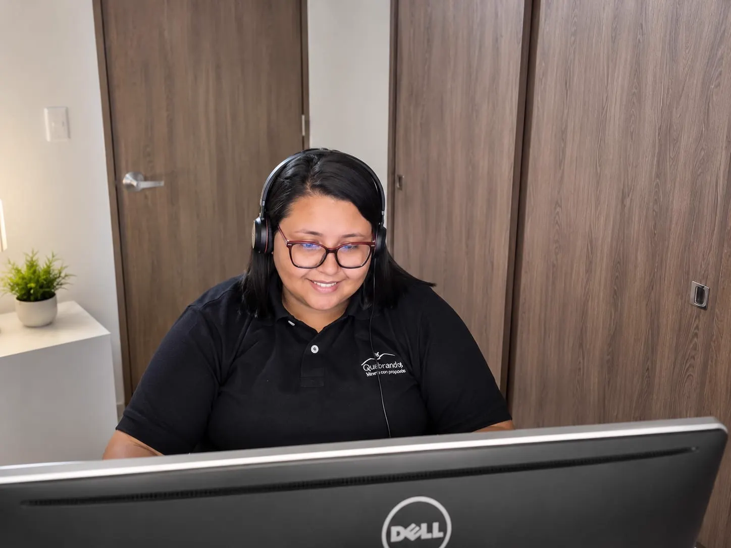 Juliana Giraldo, con diadema de audífonos, gafas y camiseta negra de Quebradona, sonríe frente a un computador en una oficina.