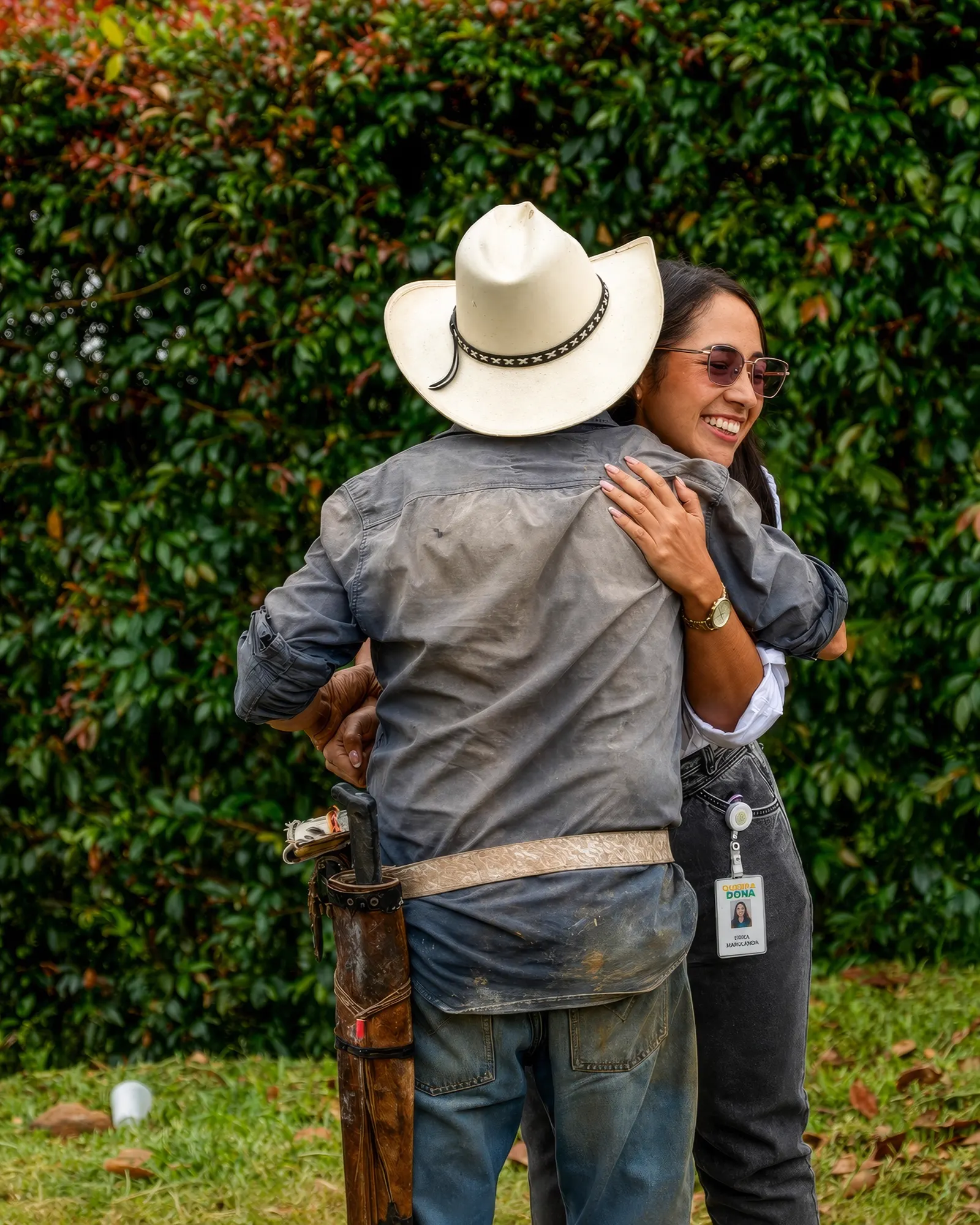Un hombre de sombrero blanco, camisa de trabajo y machete al cinto abraza a una mujer sonriente con gafas de sol y carné de Quebradona, frente a un seto verde.