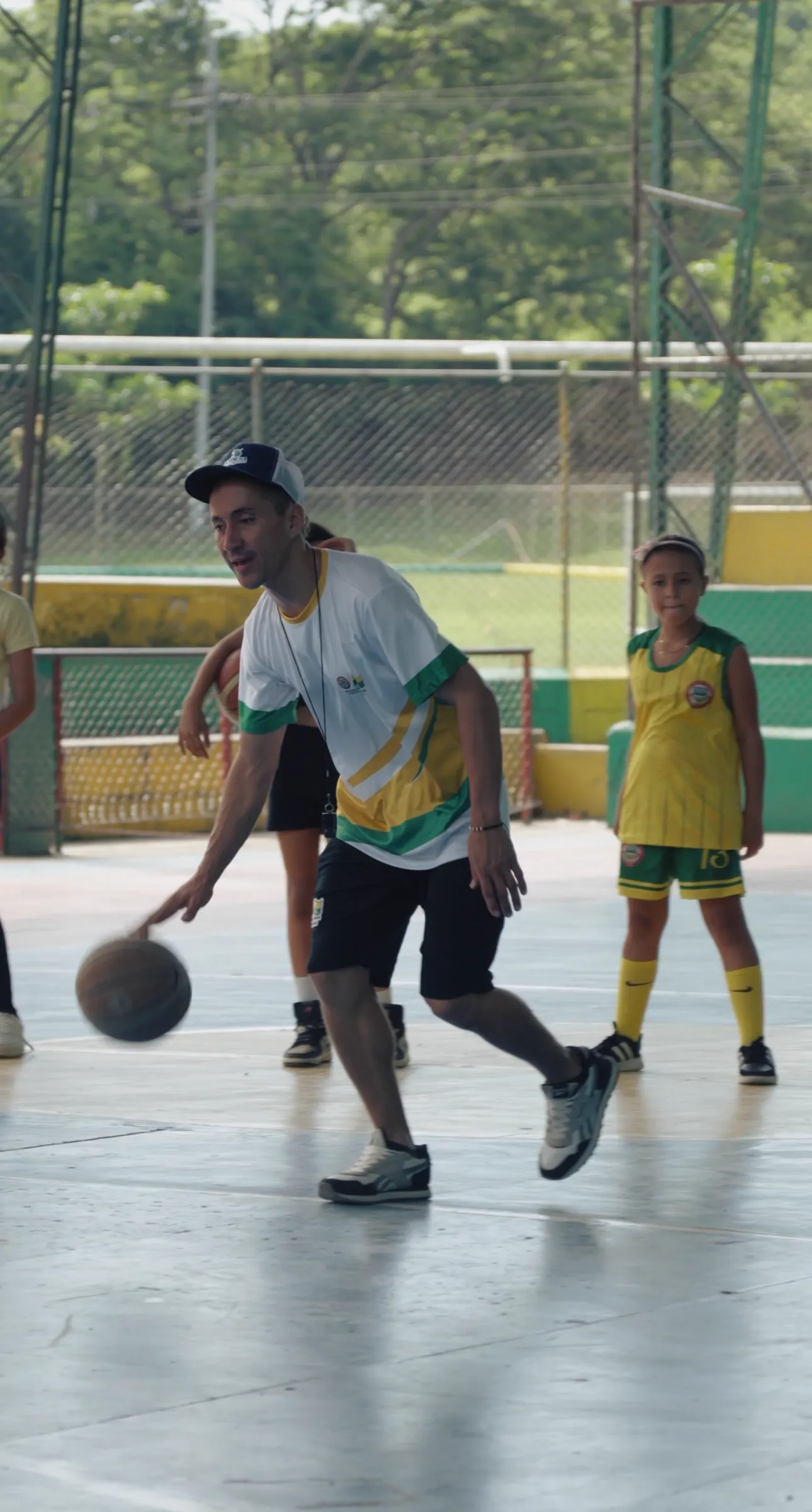 Yonifer Arango, con gorra y camiseta deportiva blanca y verde, dribla un balón en una cancha mientras una niña con uniforme amarillo observa.