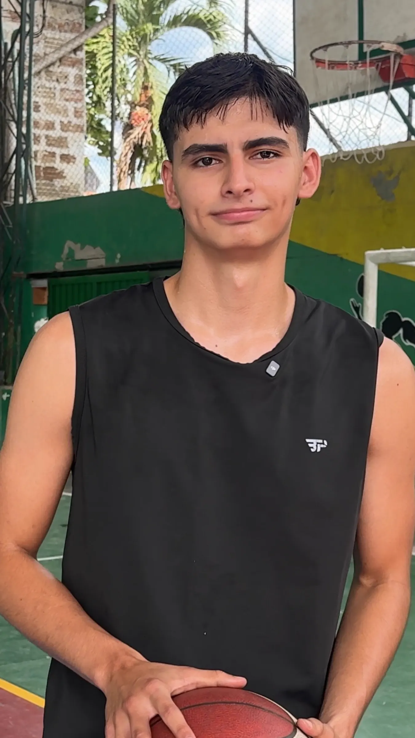 Retrato de Yosi Steven Alzate, joven de cabello oscuro con camiseta negra sin mangas, en una cancha cubierta con un aro de baloncesto detrás.