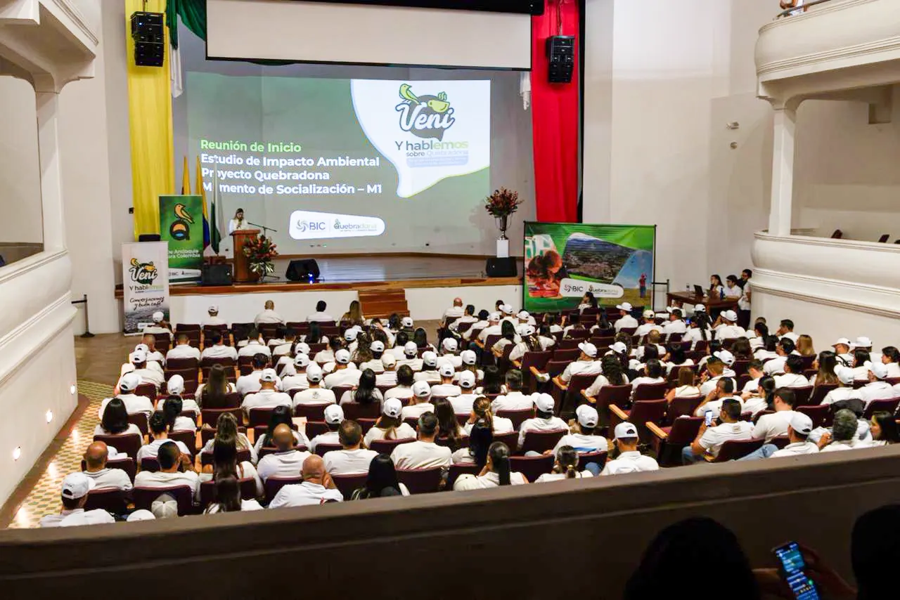 Auditorio lleno de asistentes con camisas y gorras blancas frente a una tarima; en la pantalla se lee «Reunión de inicio, Estudio de Impacto Ambiental, Proyecto Quebradona, Momento de Socialización – M1».