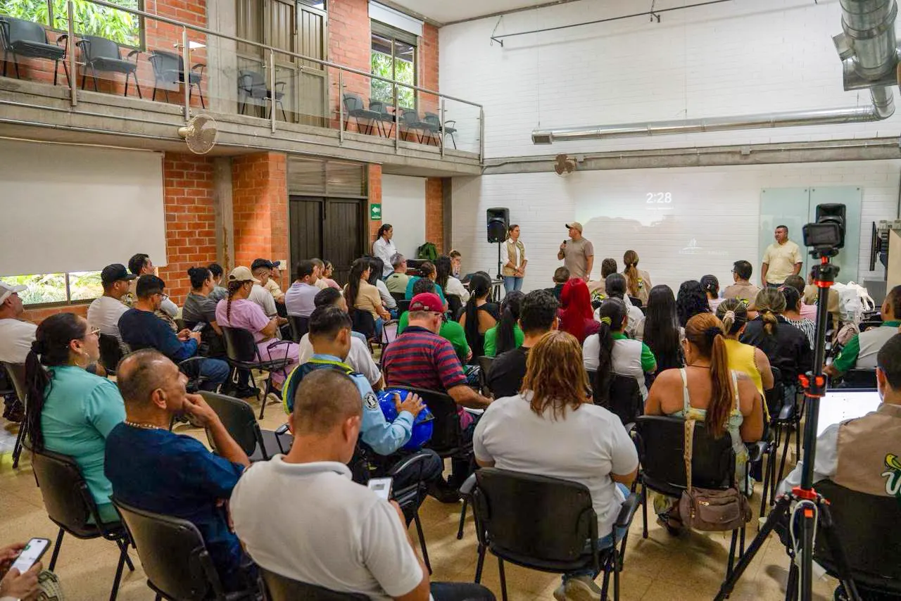 Asistentes sentados en sillas en un salón escuchan a dos expositores durante una reunión de socialización.