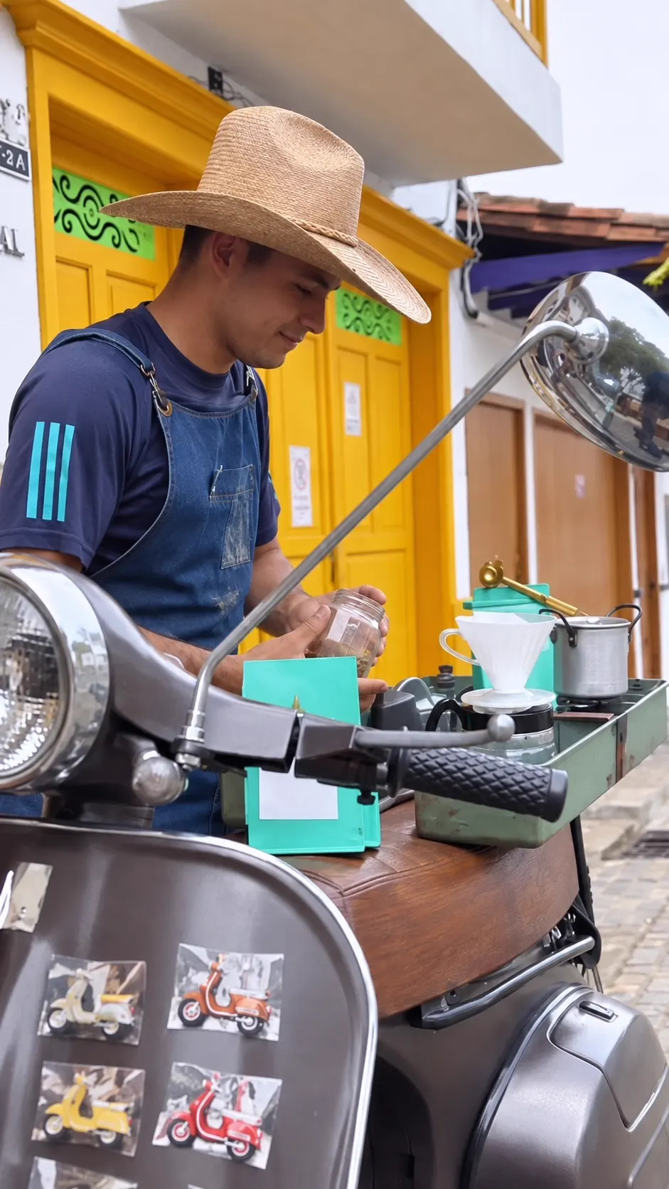 Juan Camilo Ramírez, con sombrero y delantal, prepara café en un puesto montado sobre una motoneta, frente a una fachada de puertas amarillas.