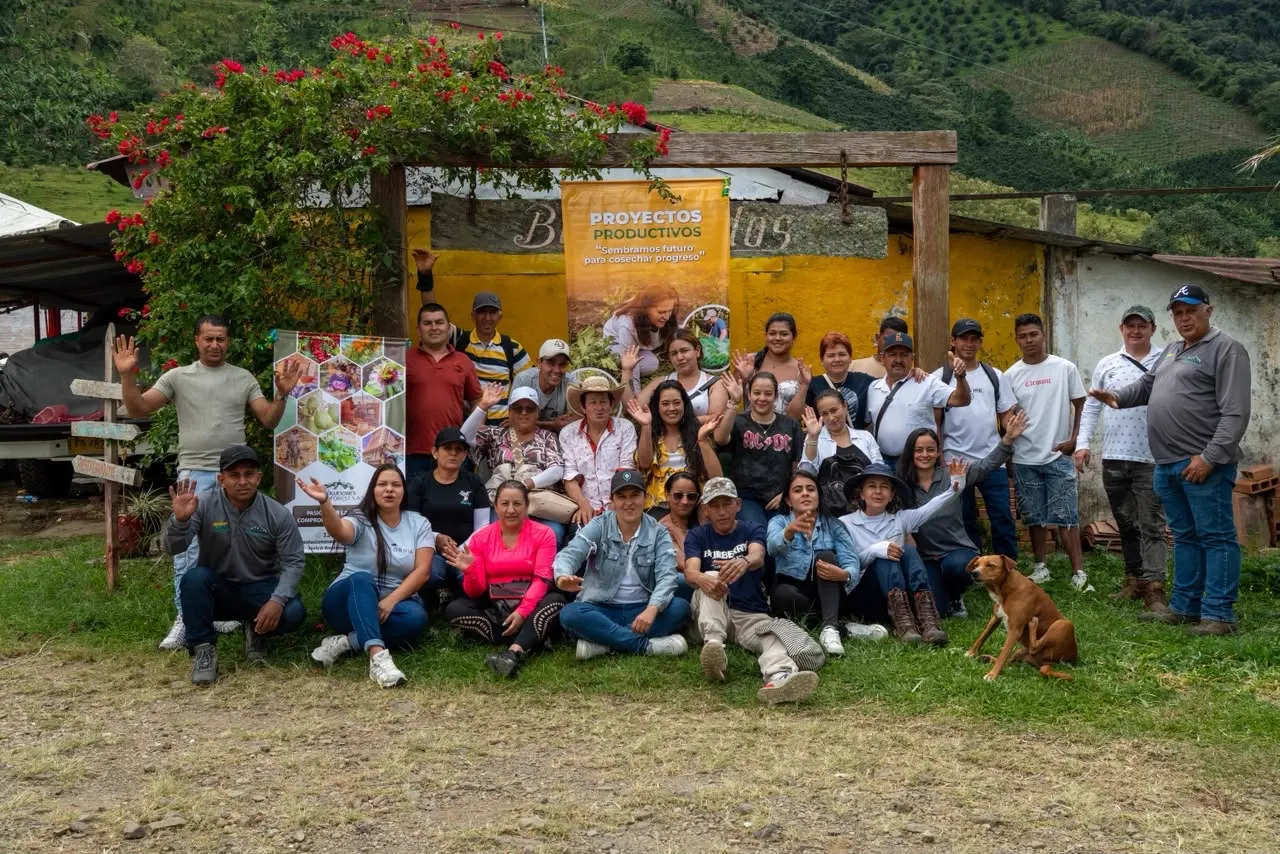 Grupo de participantes del programa posa sonriente frente a una casa campesina con un pendón de Proyectos Productivos; un perro está sentado a un lado.