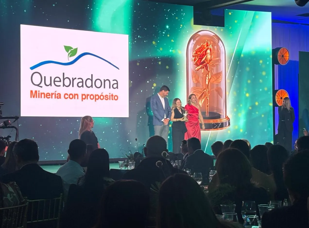 Tres personas reciben un premio en un escenario junto a una gran rosa dorada dentro de una urna de cristal; en la pantalla se ve el logo de Quebradona.