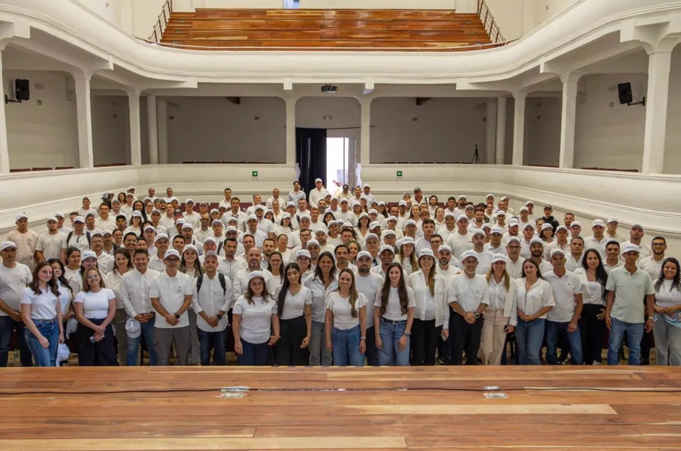 Foto grupal de cientos de personas con camisas y gorras blancas en un teatro con balcones.
