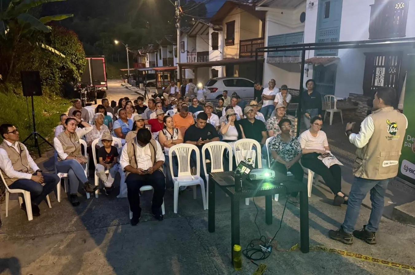 Reunión nocturna al aire libre en una calle; asistentes sentados en sillas plásticas escuchan a un expositor con chaleco de «Vení y Hablemos» junto a un proyector.