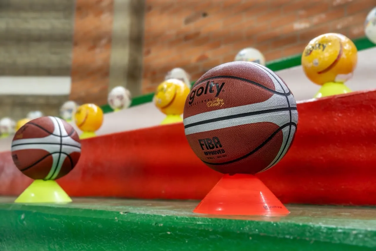 Balón de baloncesto sobre un cono naranja en unas graderías pintadas de rojo, blanco y verde, con más balones al fondo.
